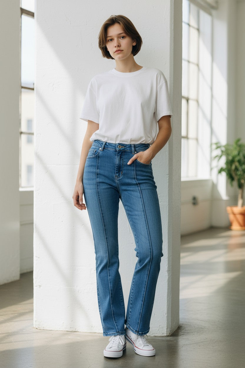 Person wearing a white t-shirt and blue jeans standing in a bright room with large windows.