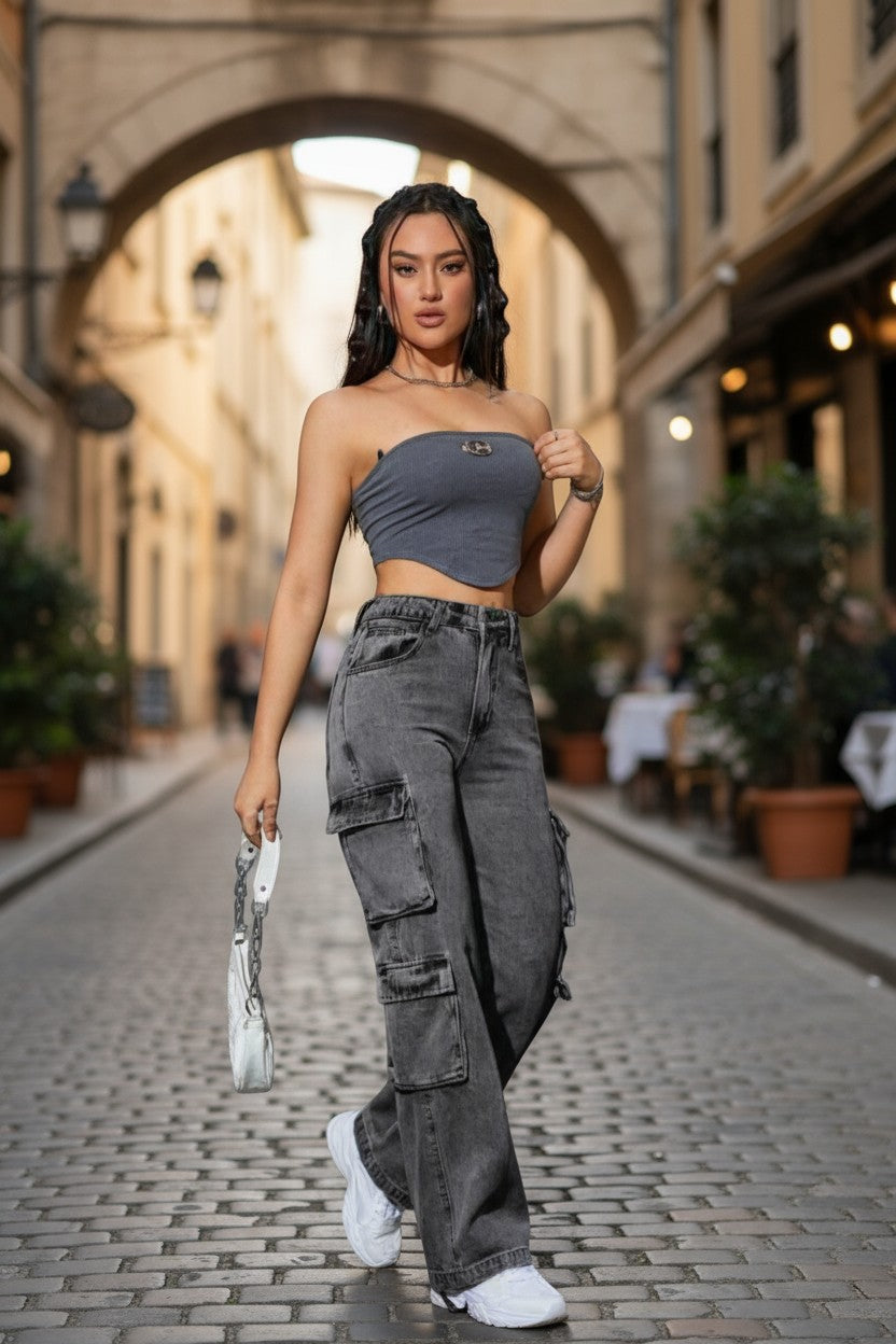 Woman in a gray strapless top and cargo pants standing on a street with arches and plants in the background.