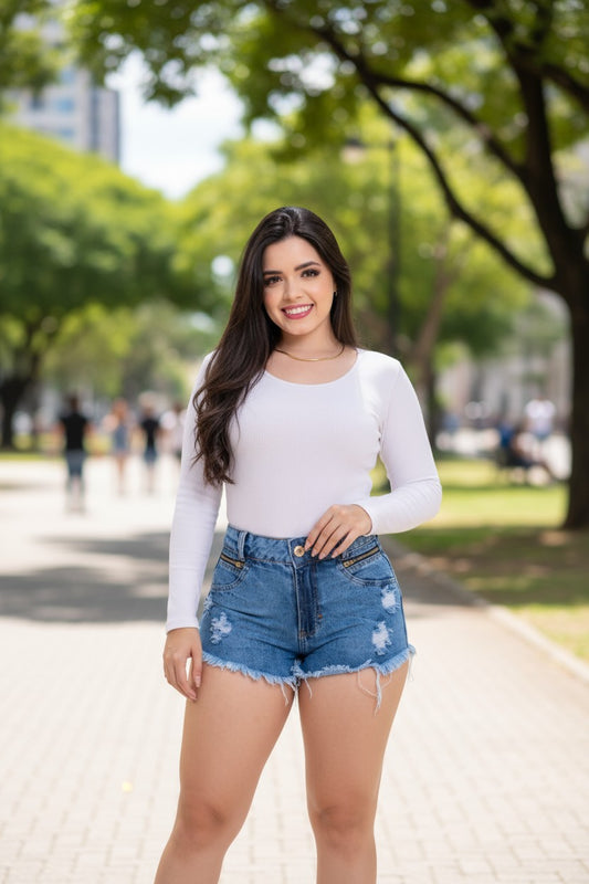 Woman in a white long-sleeve top and denim shorts standing outdoors with trees and people in the background.