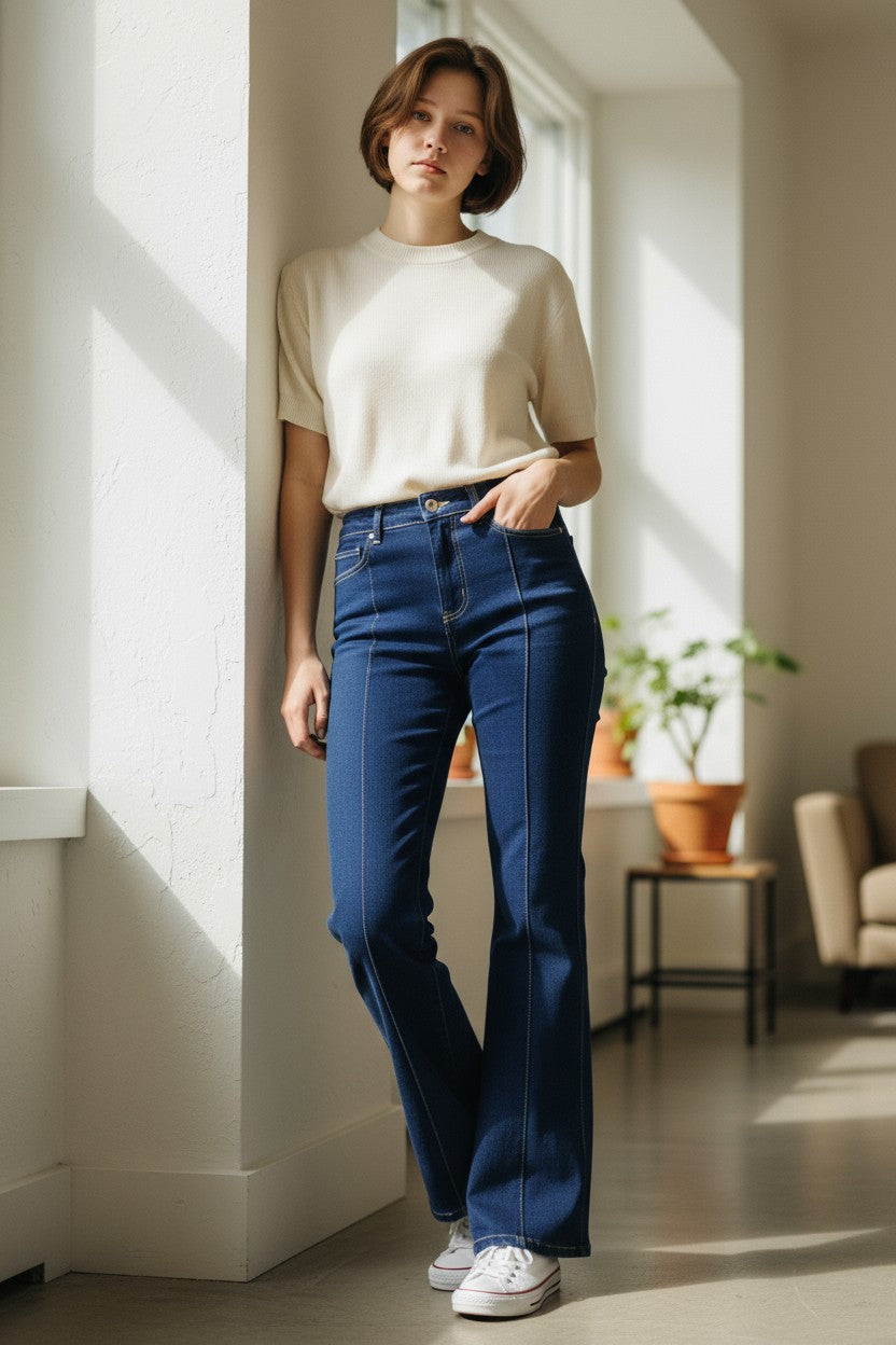 Woman wearing a beige top and blue jeans standing in a sunlit room.
